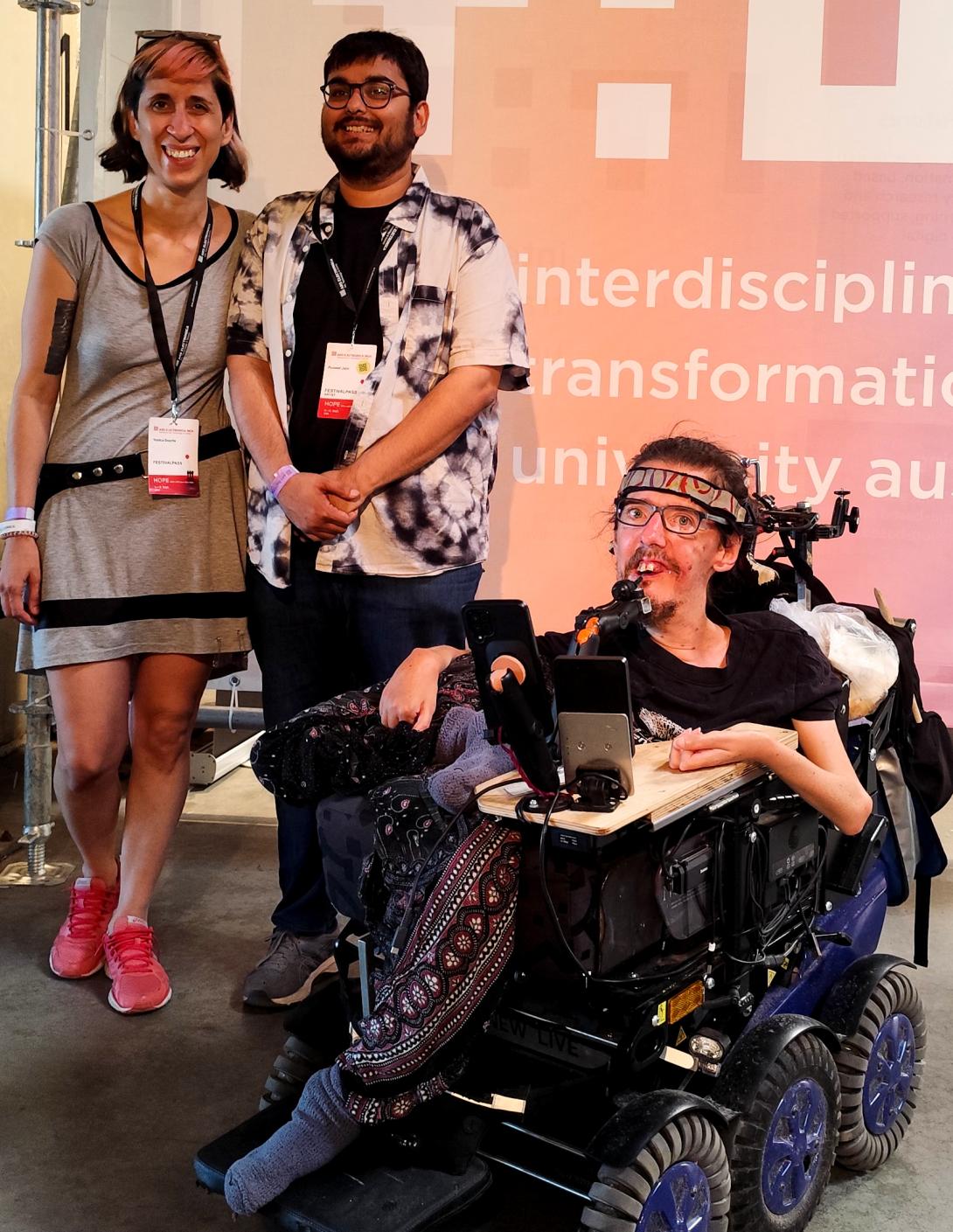 Group photo: A person using a power wheelchair with a mounted mouth joystick sits in front/right. Two people stand behind/left, wearing casual attire and event badges. All are smiling before a pink-orange event banner.
