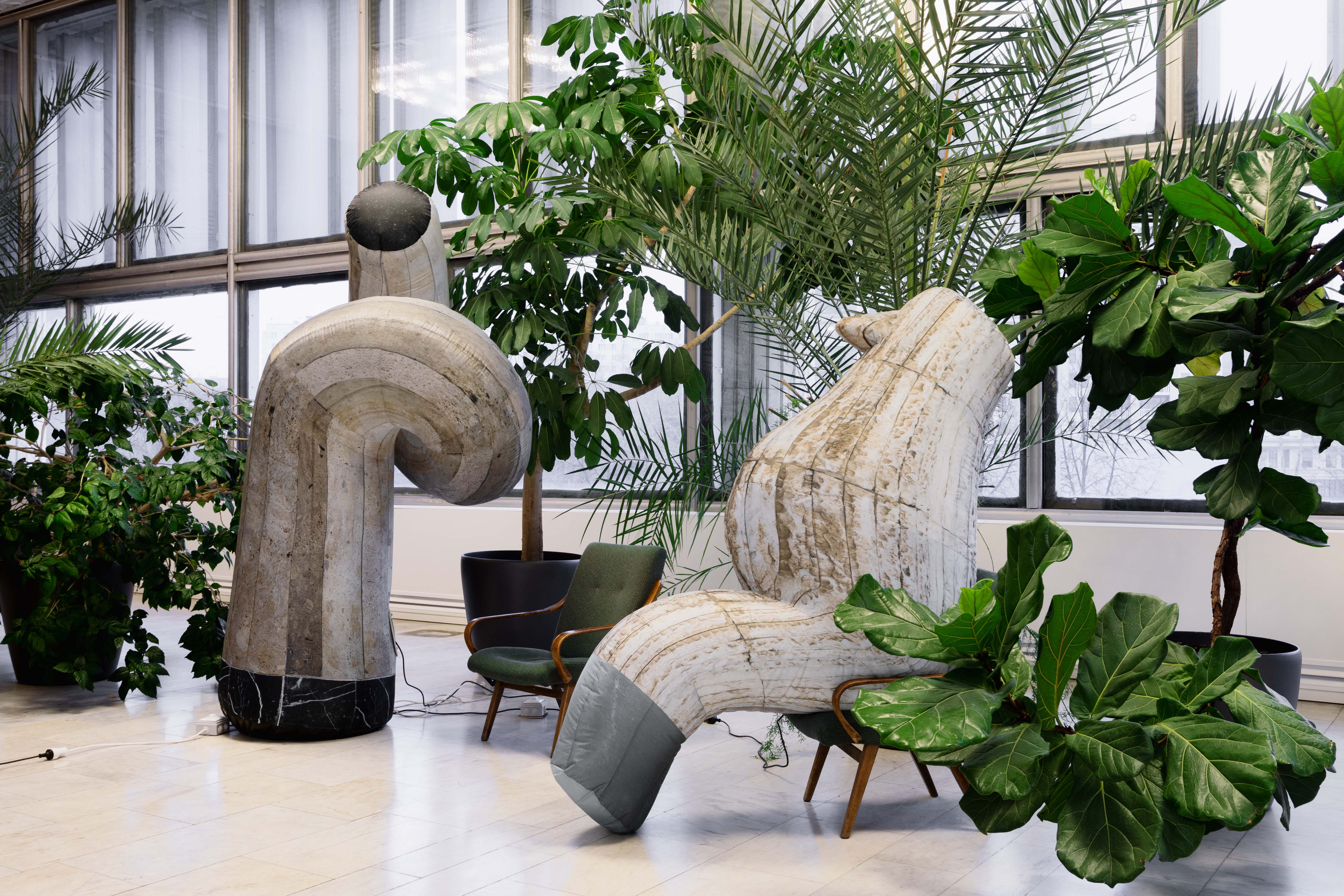 Image of a contemporary art piece surrounded by plants in the Yerevan Botanical Garden building. The art piece consists of two blown-up shapes with a light-colored, striped pattern; the one on the left is standing with a curved in its structure, the one o