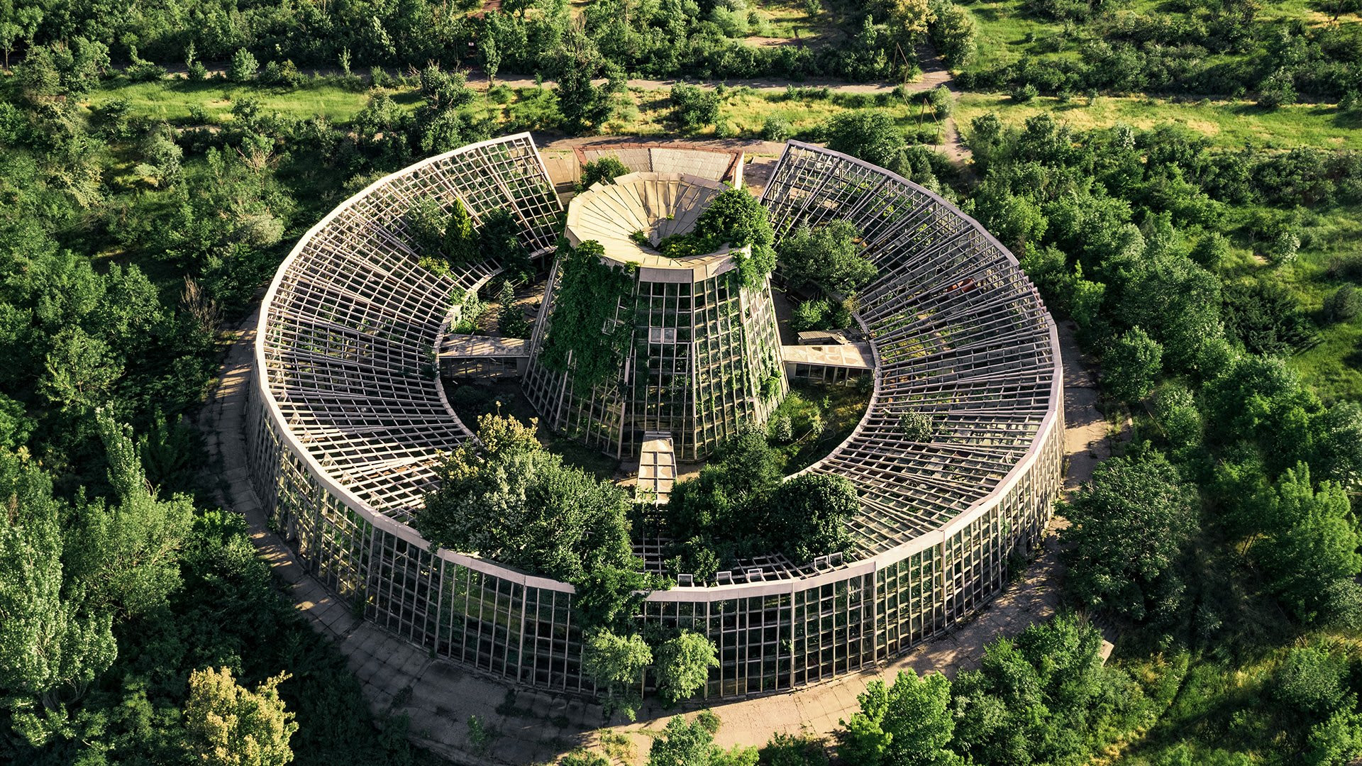 Image of an aerial view of the Yerevan Botanical Gardens. Surrounding the building is a forest of trees. The building itself is circular with a center tower. All sides show vegetation growing out of it as a sanctuary for rare plants.