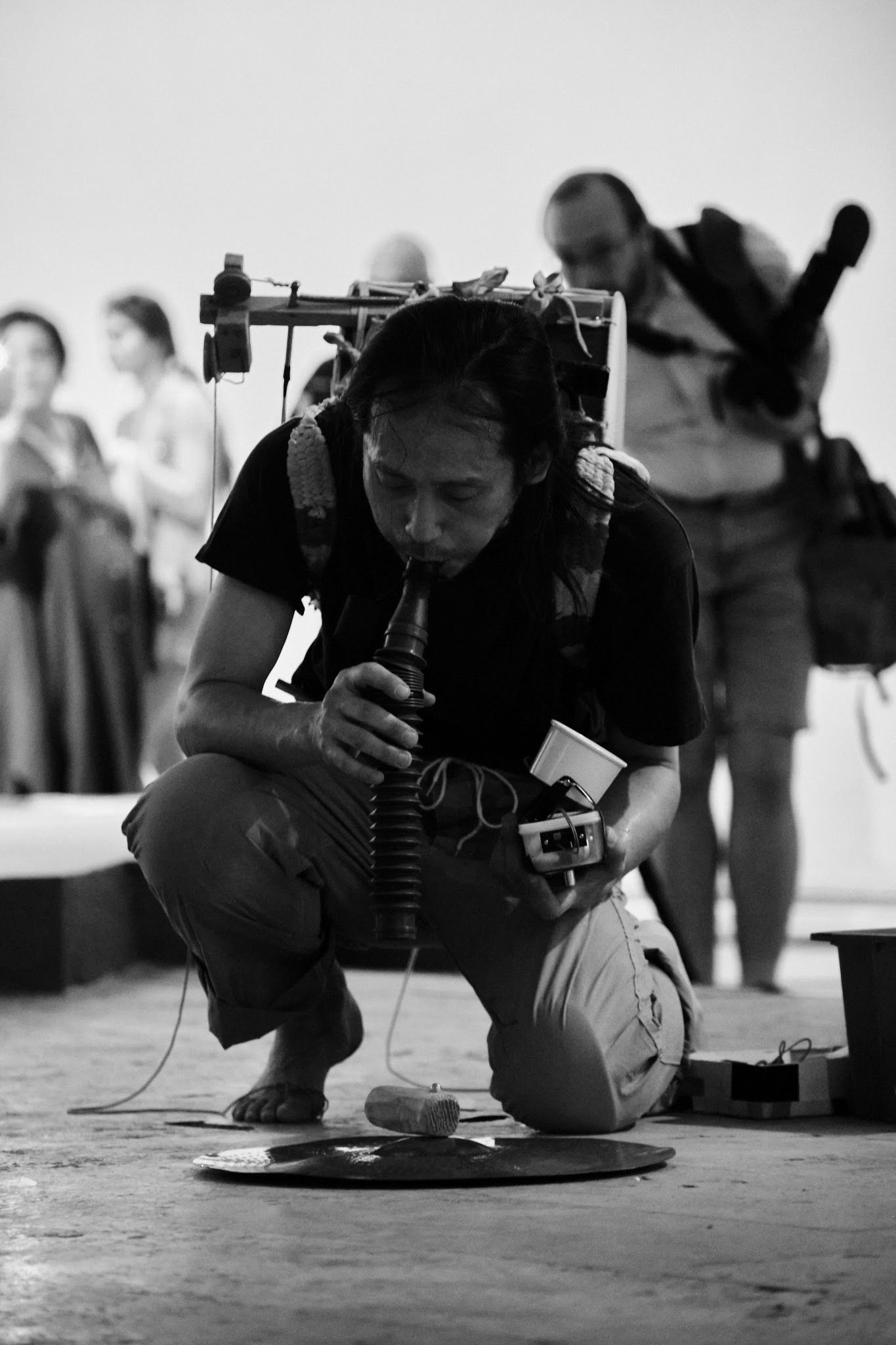 Black and white image of Nao Nishihara's sound performance in Yerevan. His performance is an abstract musical expression in a friendly street style with a drum on his back.
