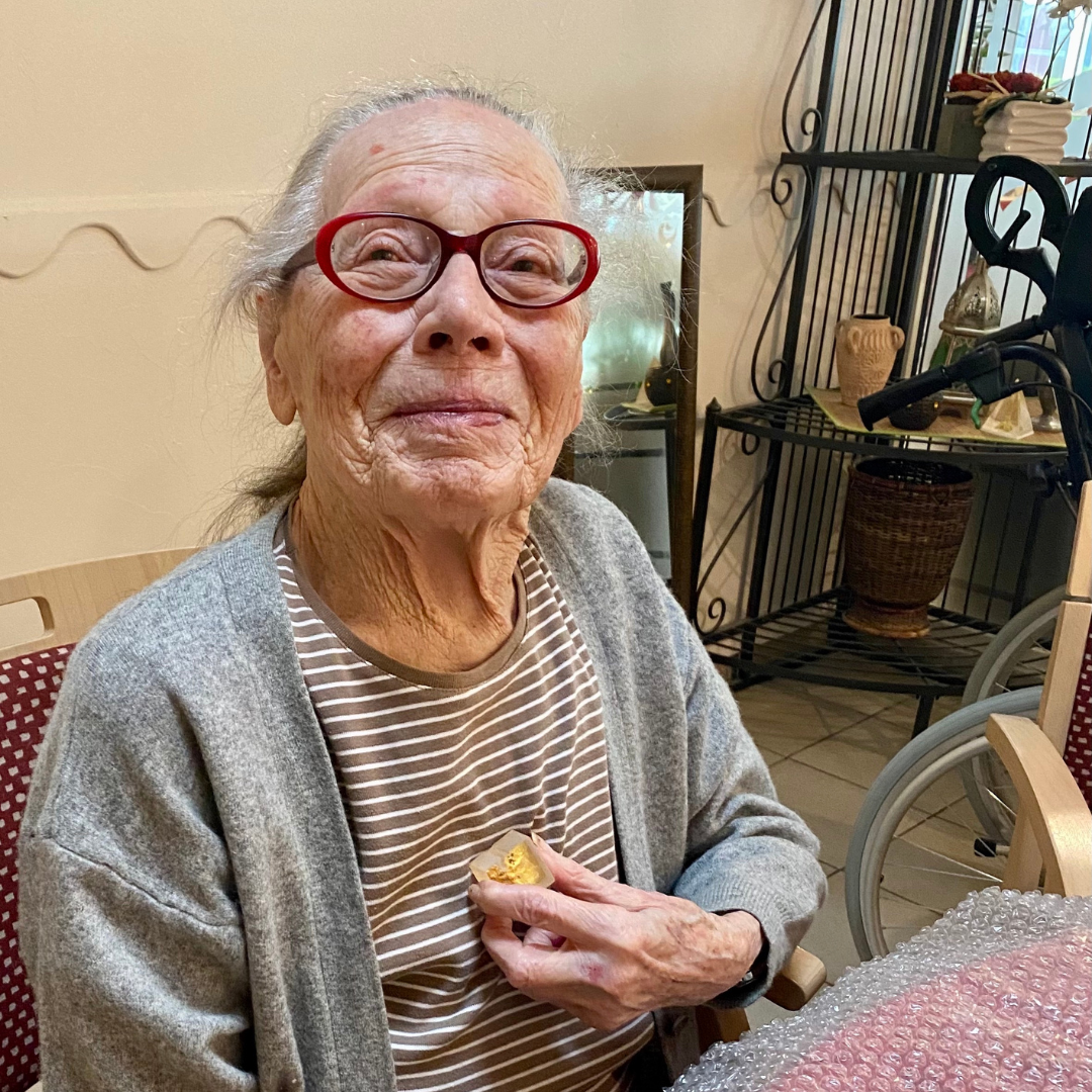 A portrait of Vera Molnár, Paris 2021. She’s smiling with frazzled white hair, wearing red glasses, and holding a small crystal-like object to her chest. Image courtesy of Francesca Franco.
