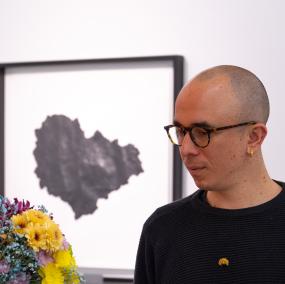 A bald individual with pale skin tone stares at a mixed bouquet of flowers. They are wearing a black sweater and has circular glasses on. In the background is a blurry painting of a seashell in black and white. 
