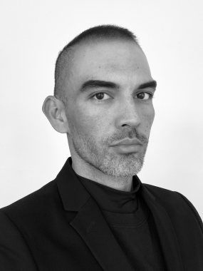 Black and white image of a masculine presenting individual. They have short hair, almost in a buzzcut and a scruffy beard. They are wearing a black suit jacket and a matching turtleneck sweater. 
