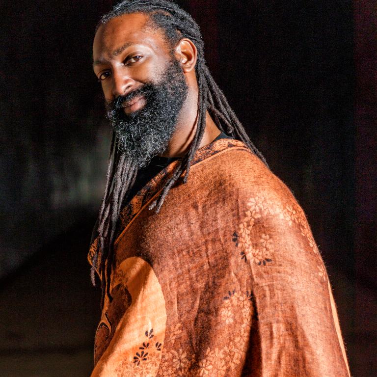 Mr Hunter, A Black and Indigenous person with dark chocolate skin from his mother. He has almond shaped eyes with long lashes. He has long black dreadlocks tied in a low braid and a full beard. Antoine is wearing a brown top and smiling.
