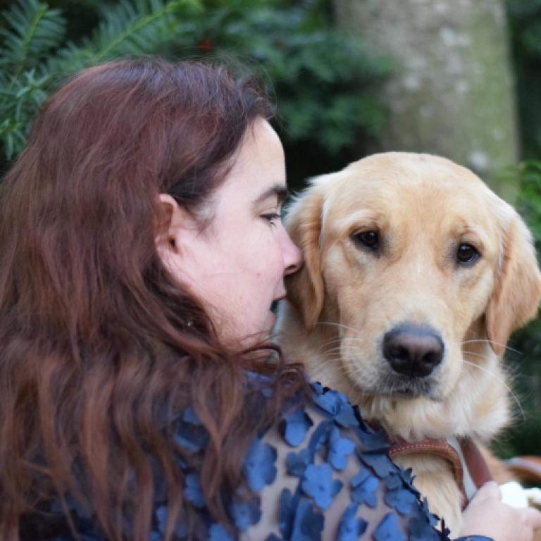 Caucasian fat woman with long auburn hair wearing a sheer top covered in navy tactile flowers in profile facing her golden retriever guide dog with bright eyes and a nearly heart shaped nose
