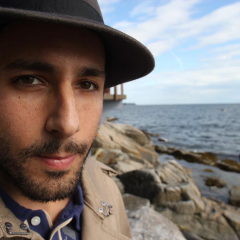 Against a blurry background of rocks and ocean, a close-up of an olive-skinned man with brown eyes, a dark, close-trimmed beard, and a gray hat.