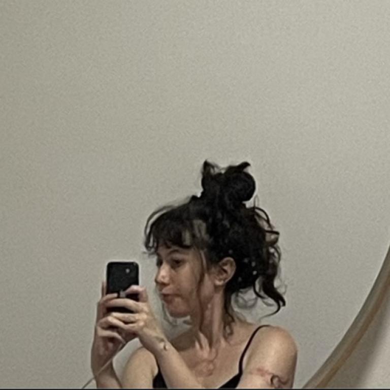 A mirror selfie of Meesh. She has brown curly hair tied up in a messy bun and is holding her phone with both hands.