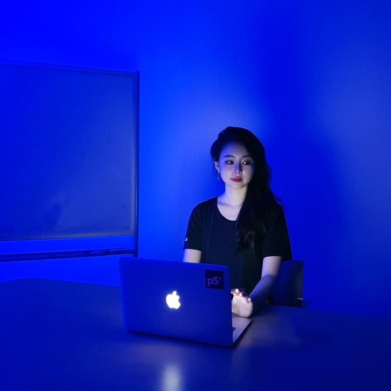 A person with black long hair in black t-shirt is sitting in front of a laptop at a desk, in a room projected with bluescreen.
