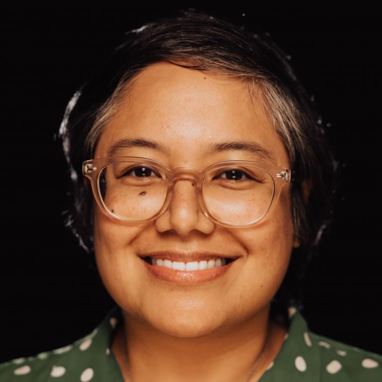 A brown skinned filipino person with clear glasses, freckles, short dark hair wearing a green and white polka dot button up.