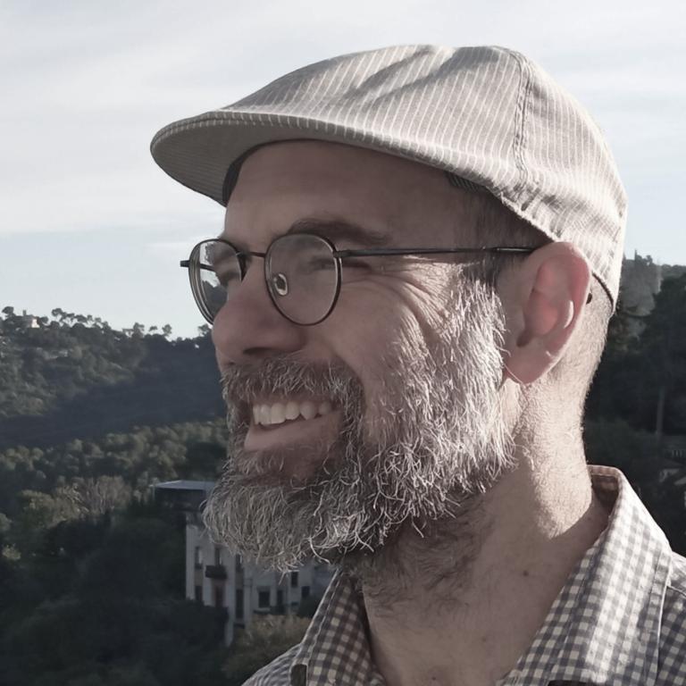 50-year old white male with grayish beard, glasses and hat looking left against a green mountain background