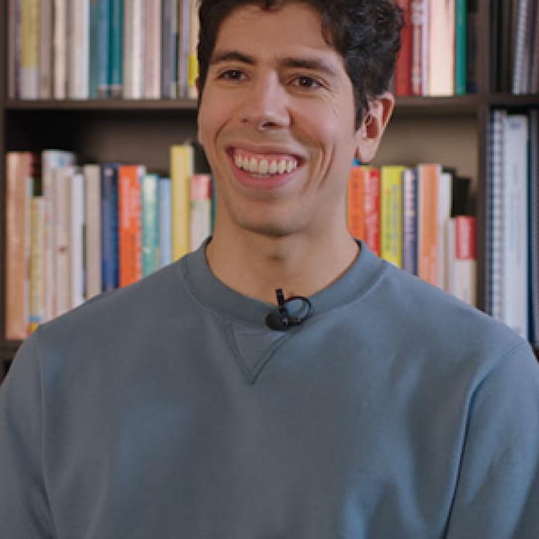 A male-presenting, half-Latino person wearing a blue sweater smiles.