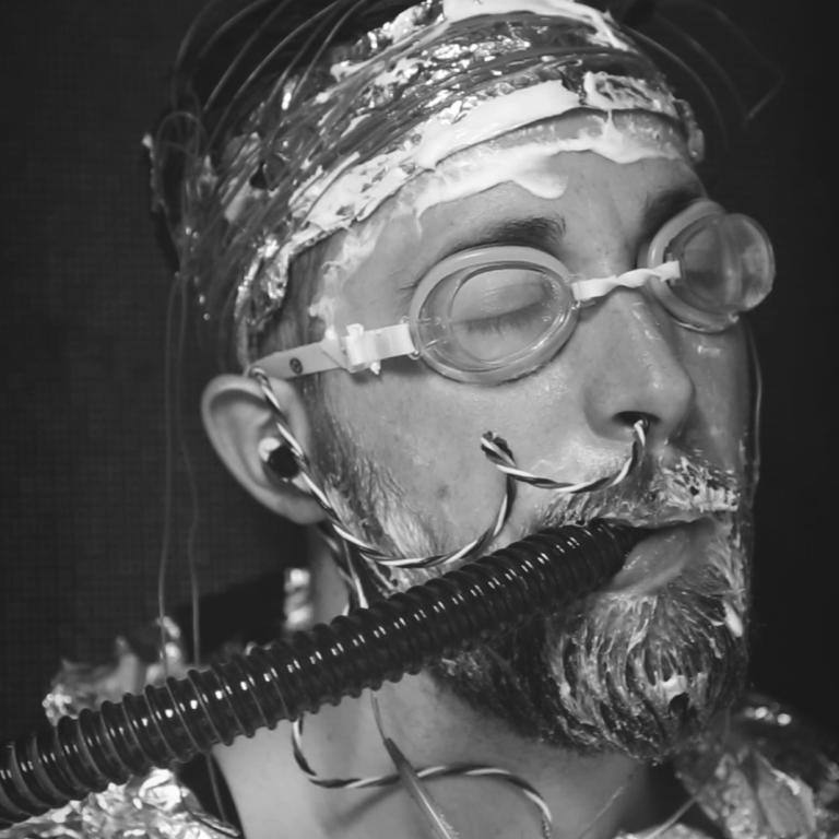 A black-and-white photo shows a man with closed eyes, wearing goggles and tin foil, breathing through a tube in his mouth.